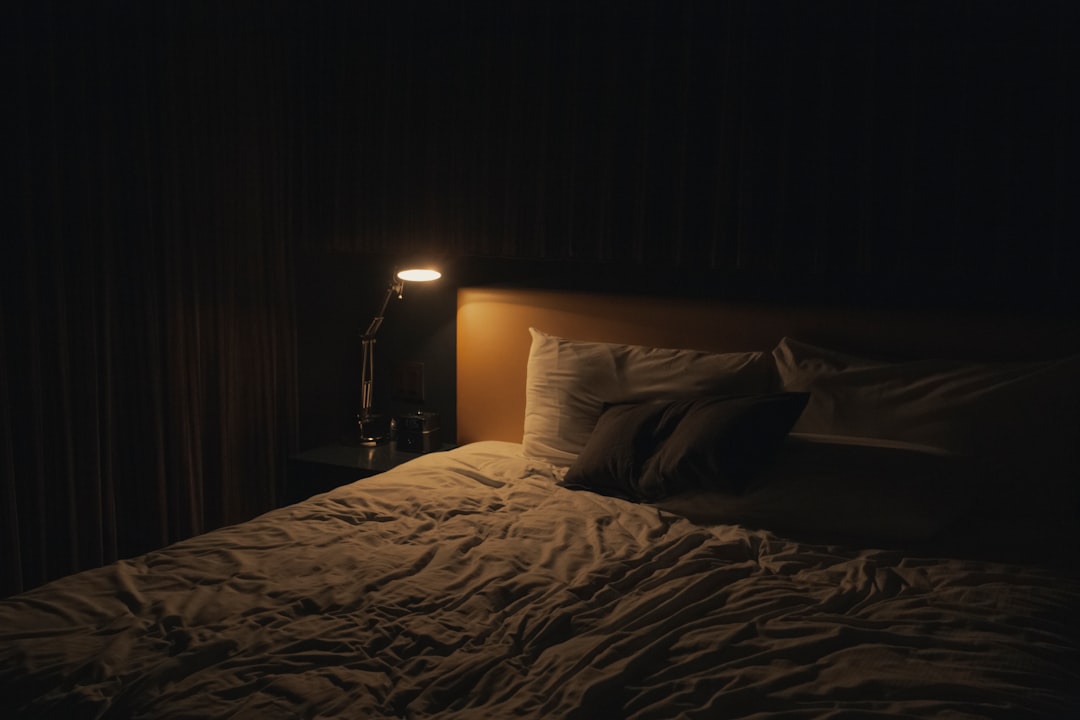 Warm evening light on a nightstand with a glass of water, softgel capsules, and a dim lamp beside a made bed